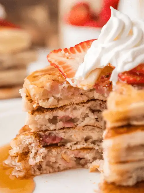 Stack of fluffy easy strawberry pancakes cut open to show fresh strawberry pieces inside, topped with whipped cream and maple syrup — a simple fresh strawberry brunch recipe.