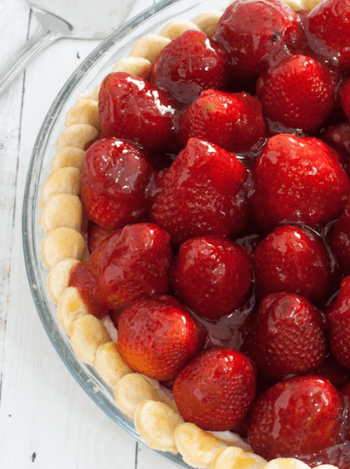 Overhead close-up of a fresh strawberry pie in a glass pie dish packed with whole glazed strawberries in a shiny red sauce nestled in a golden fluted crust — a classic strawberry dessert recipe that puts peak season berries front and center.