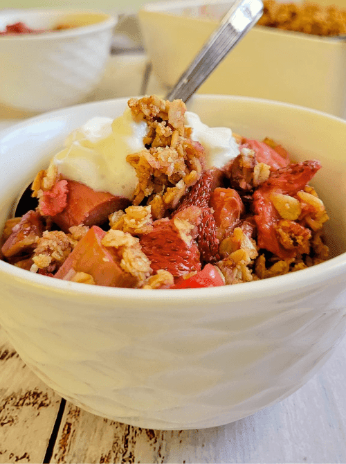 A white bowl of strawberry rhubarb crisp topped with a dollop of whipped cream and a golden oat crumble, with additional servings visible in the background — one of the best things to make with strawberries all summer long.