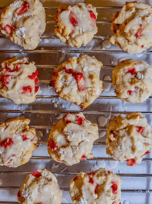 Overhead view of strawberry scone cookies with lemon icing drizzled over fresh strawberry pieces cooling on a wire rack — an easy drop-style fresh strawberry brunch recipe.