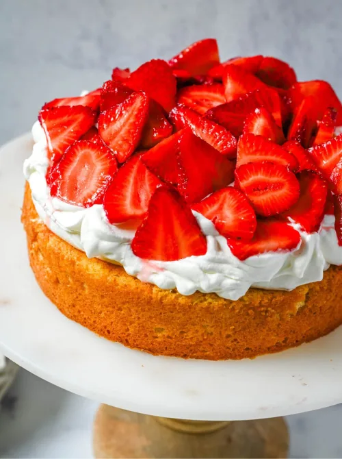 Single layer strawberry shortcake cake on a marble cake stand piled high with macerated fresh strawberries and whipped cream over a golden buttermilk sponge — a from-scratch homemade strawberry cake recipe with classic shortcake flavors in every slice
