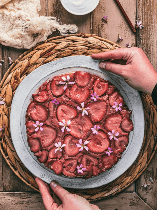 Overhead view of dairy-free strawberry upside down cake with caramelized strawberry slices and purple edible flowers on a rustic gray plate, hands holding the plate over a woven charger — a stunning from-scratch strawberry cake recipe with zero decorating required