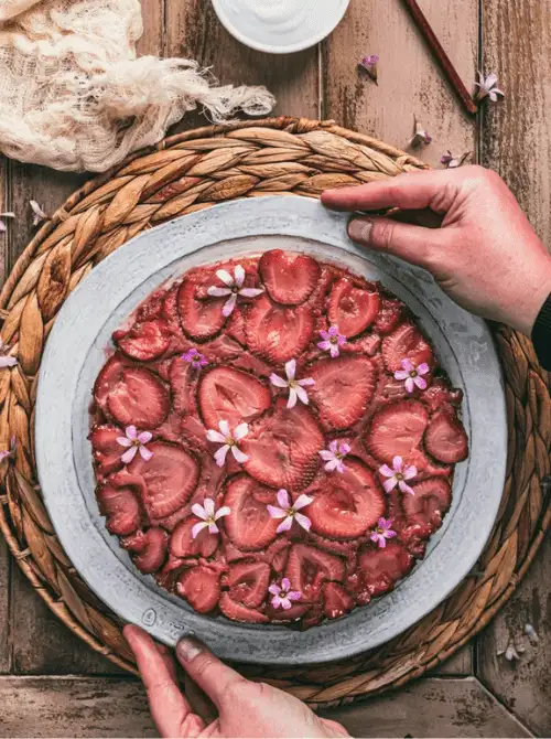 Overhead view of dairy-free strawberry upside down cake with caramelized strawberry slices and purple edible flowers on a rustic gray plate, hands holding the plate over a woven charger — a stunning from-scratch strawberry cake recipe with zero decorating required