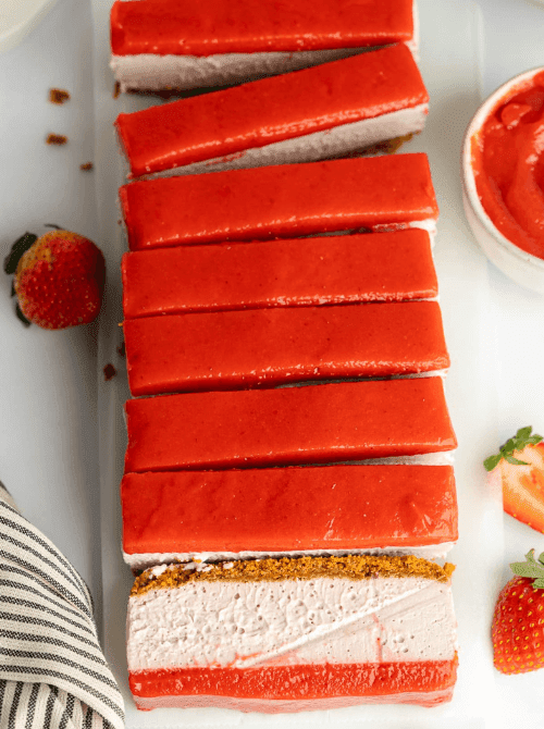 Sliced vegan strawberry cheesecake on a white serving board showing layers of biscoff cookie crust, pink strawberry cream filling, and a glossy strawberry curd topping, with fresh strawberries alongside — a stunning no-bake summer strawberry dessert that happens to be completely dairy-free.