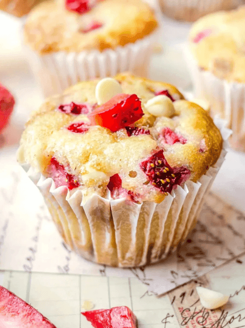 Close-up of a domed white chocolate and strawberry muffin in a paper liner with visible strawberry pieces and white chocolate chips on top — a bakery-style fresh strawberry brunch recipe.